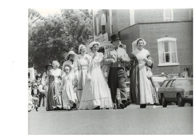 Group of woman and girls in C19th period dress, led by two women arm in arm with a gentleman in C19th frock coat and top hat, walking past a brick building.