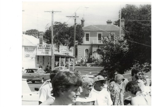 Down street, crowd close up at front, shop on left, old 2-storey bank centre, trees to the left.