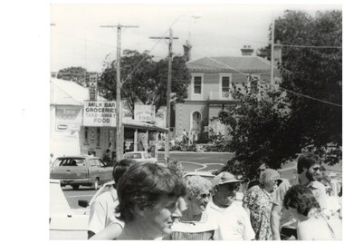Down street, crowd close up at front, shop on left, old 2-storey bank centre, trees to the left.