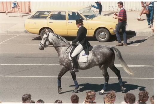 Lady in full dressage outfit, riding dappled greay horse with plaited mane, past yellow station wagon.