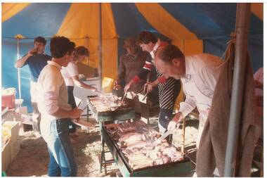 Sausage sizzle in yellow and blue tent, four men and two women stand either side of wood-fire barbecues cooking meat and sausages.