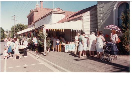 Buildings on right, rendered bank, wooden old store with veranda, two-story brickbrick, parking spaces on left, stalls and crowds on foorpath and uinder veranda.