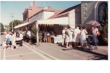 Buildings on right, rendered bank, wooden old store with veranda, two-story brickbrick, parking spaces on left, stalls and crowds on foorpath and uinder veranda.
