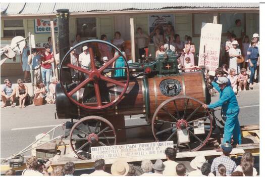 Old wood, brass and iron wagon-mounted 1905 steam engine in procession passing crowds and shops.