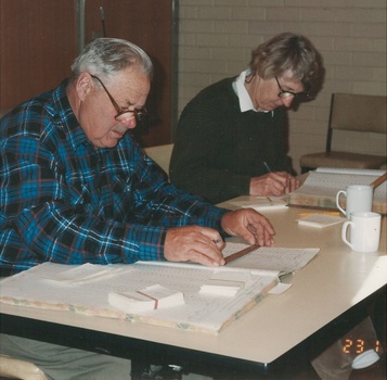 Man in check shirt, and lady in jumper, both wear glasses, sitting at table indexing rate books, books, papers and mugs of coffee on the table.