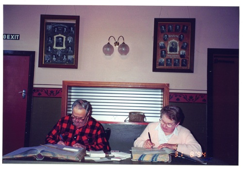 Man in check shirt and woman in cardigan sitting at table indexing old large rate books, large collage photos on wall behind. 