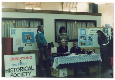 Display table with signage boards, four volunteers at table, two sitting, two standing, Inmside large weatherboard lined hall.