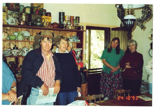 Volunteers stand around table in cottage kitchen, shelving cabinet behind them, filled with kitchenware, crockery etc.