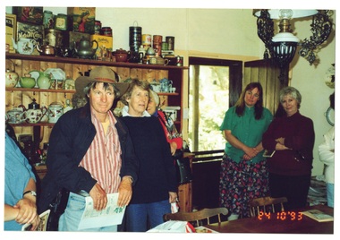 Volunteers stand around table in cottage kitchen, shelving cabinet behind them, filled with kitchenware, crockery etc.
