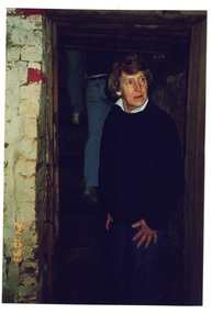Middle-aged lady in dark jumper standing in gloomy doorway of painted brick cellar.