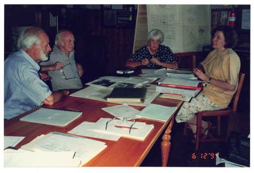 Two men and two women sitting at wooden table, table covered with documents and books.