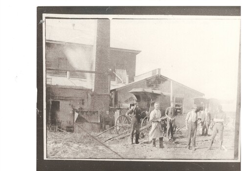 Group of workmen in front of two story concrete block industrial building, tall brick chimney, 