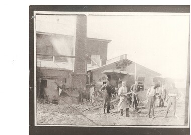 Group of workmen in front of two story concrete block industrial building, tall brick chimney, 