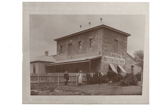 Two story concrete block factory, weatherboard extension on left connected to iron-roofed veranda, picket fence, four men standing in front. Buninyong Butter Factory sign painted on end wall.