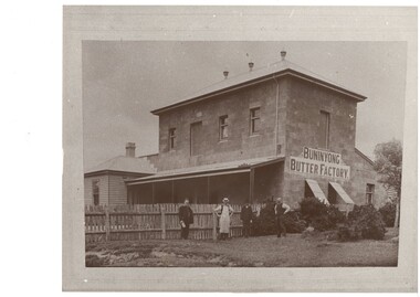 Two story concrete block factory, weatherboard extension on left connected to iron-roofed veranda, picket fence, four men standing in front. Buninyong Butter Factory sign painted on end wall.