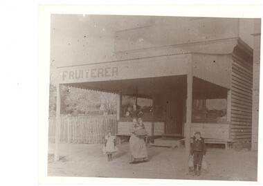 Black and white photograph of the exterior of Butcher's Fruiterer Shop featuring a woman holding a child and two other children standing in the foreground