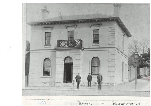 Two story bank building, shallow entrance porch with balcony over arched double doors, arched window either side, rectangular windows upper floor. Three men in front.