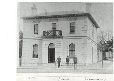 Two story bank building, shallow entrance porch with balcony over arched double doors, arched window either side, rectangular windows upper floor. Three men in front.