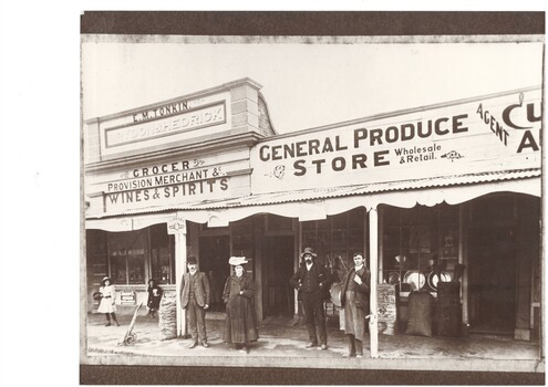Two late Victorian shop-fronts, a grocer and a Produce store, with veranda, assorted people in Edwardian clothes, some produce on display.