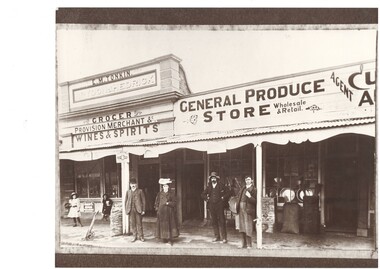Two late Victorian shop-fronts, a grocer and a Produce store, with veranda, assorted people in Edwardian clothes, some produce on display.