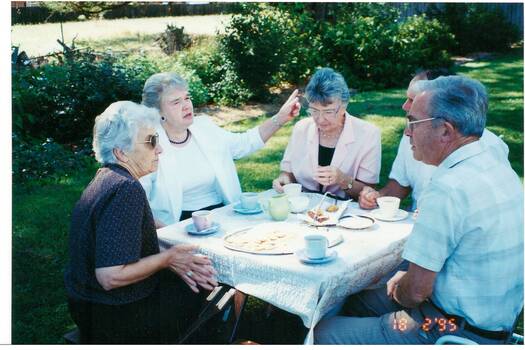 Three middle-aged ladies and two men in casual clothes, seated in a garden at a square picnic table with cups of tea, a cake and biscuits.