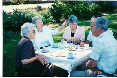 Three middle-aged ladies and two men in casual clothes, seated in a garden at a square picnic table with cups of tea, a cake and biscuits.