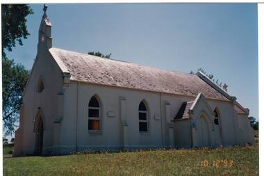 Corner view of rendered Victorian era Church, slate roof, porch entrance and pointed arch windows on side, door, small rose window and small open bell tower .at end,