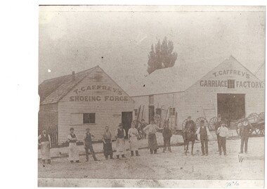 Faded B&W photo two simple wooden buildings, peaked shingle roofs, on left a shoeing forge with small door and window, on right  a carriage factory with single large open doorway, carriage wheels and shafts lean against the walls, a line of 12 men and two horses, and one boy on one horse, on the road in front.