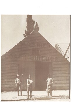 Simple wooden factory, high peaked roof, large double doors, one open, three men in front wearing work-clothes, one with leather apron.
