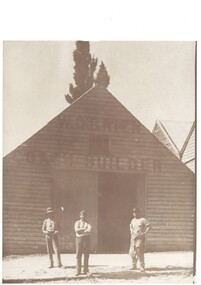 Simple wooden factory, high peaked roof, large double doors, one open, three men in front wearing work-clothes, one with leather apron.