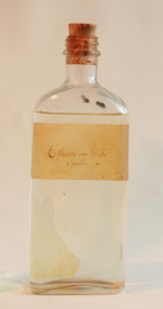 Ether was once stored in clear glass bottles for easy transportation. A discoloured white label has "Ether in Oil" handwritten on it, and the bottle has a cork stopper.