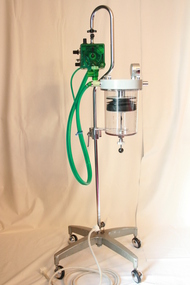 Attached to an upright pole, the four metal casters make this apparatus portable. The green tubing is indicative of a Bird Ventillator. 