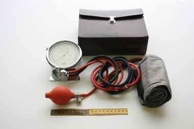 Long strands of black and red rubber tubing, are attached to a gauge at one end, and a fabric cuff at the other, with a leather carry bag sitting behind them.