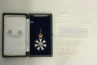 Nestled in a velvet and satin lined box, the medal of the Order of St Michael and St George has a round enamel centre, with with six white, enamel rays leading out from it. Atop one of the rays, is a golden coloured enamel crown, leading to a ring for attaching a ribbon.