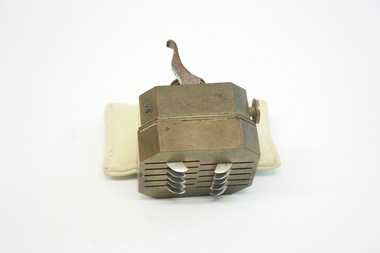 A scarificator made of brass, in a squarish shape, with a lever at the top. At the base of the object is a series of ten slits, through which metal blades emerge once the lever is pushed. 