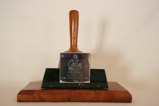 Plain highly polished wooden pestle sits on a central walnut block  on a flat wooden base, a silver plaque is attached on a piece of nephrite stone to one side of the board.