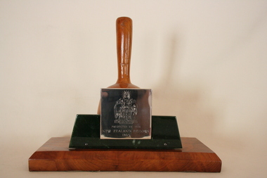 Plain highly polished wooden pestle sits on a central walnut block  on a flat wooden base, a silver plaque is attached on a piece of nephrite stone to one side of the board.