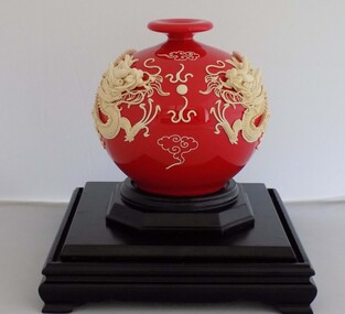 A white dragon, in raised relief, is wrapped around a round red laquered vase, resting on a double tiered wooden plinth.