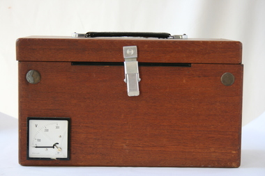 A rectangular wooden box with chrome clasp connecting the lid to the base and enabling it to be locked in place. There is a square, white faced dial, or gauge on the bottom left hand corner of the box's face and a handle lying flat against the top of the lid.