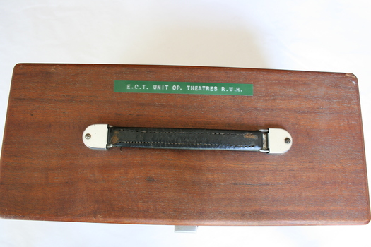 The leather strap of the machine's handle has been well-worn, signifying much use. Above the handle is a green label with white printed text in relief. The text reveals the machine to be an ECT machine and the property of the Royal Women's Hospital.