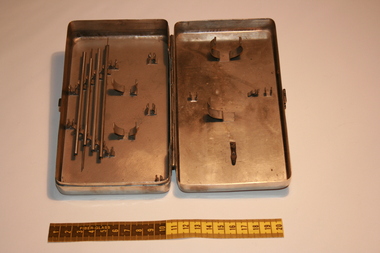 A stainless steel syringe kit sitting open on a table. There are no syringes in the container, only hollow metal rods.