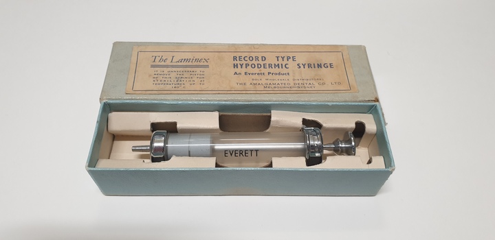 A greenish grey cardboard lies open, with its lid behind it. Inside the box is a glass barrelled syringe with a metal plunger. The manufacturer's name, Everett, is printed on to the barrel.