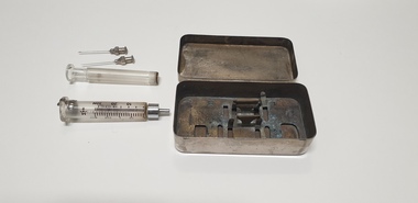 A metal box lies open on a table, with the contents around it including a glass barrel syringe and glass plunger, and two metal needles.