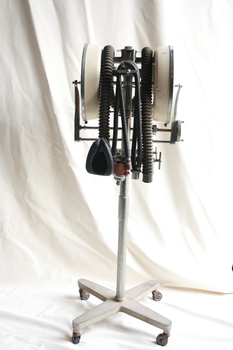 An Austox D-M gas anaesthesia machine affixed to a stand with castors. The machine has attached black corrugated tubing with masks to go over the nose and mouth. There are two white, circular chambers on either side of the machine