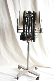 An Austox D-M gas anaesthesia machine affixed to a stand with castors. The machine has attached black corrugated tubing with masks to go over the nose and mouth. There are two white, circular chambers on either side of the machine