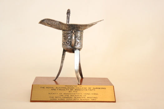 A silver tripod Chinese wine cup set on an angle-fronted wooden plinth with attached brass plaque.