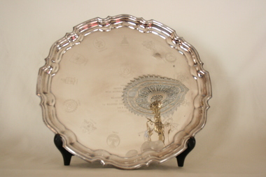 A large, round silver-plated salver with a raised geometric design around the rim. Thirteen crests have been engraved around the tray text and coat of arms.