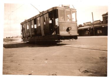 A vintage electric tram car numbered 21, with multiple windows and a dark exterior, sits on tracks on a paved street. In the background, a row of shops with awnings lines the street, including a sign for "ROODAS GROCER & PROVISION MERCHANT" and "PASTRY COOK".