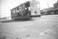 A vintage electric tram car numbered 21, with multiple windows and a dark exterior, sits on tracks on a paved street. In the background, a row of shops with awnings lines the street, including a sign for "ROODAS GROCER & PROVISION MERCHANT" and "PASTRY COOK".
