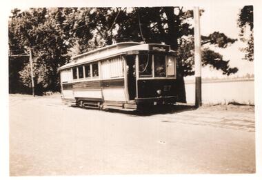 A two-colored vintage tram numbered 27 is stopped on tracks next to a road with a body of water and trees in the background.